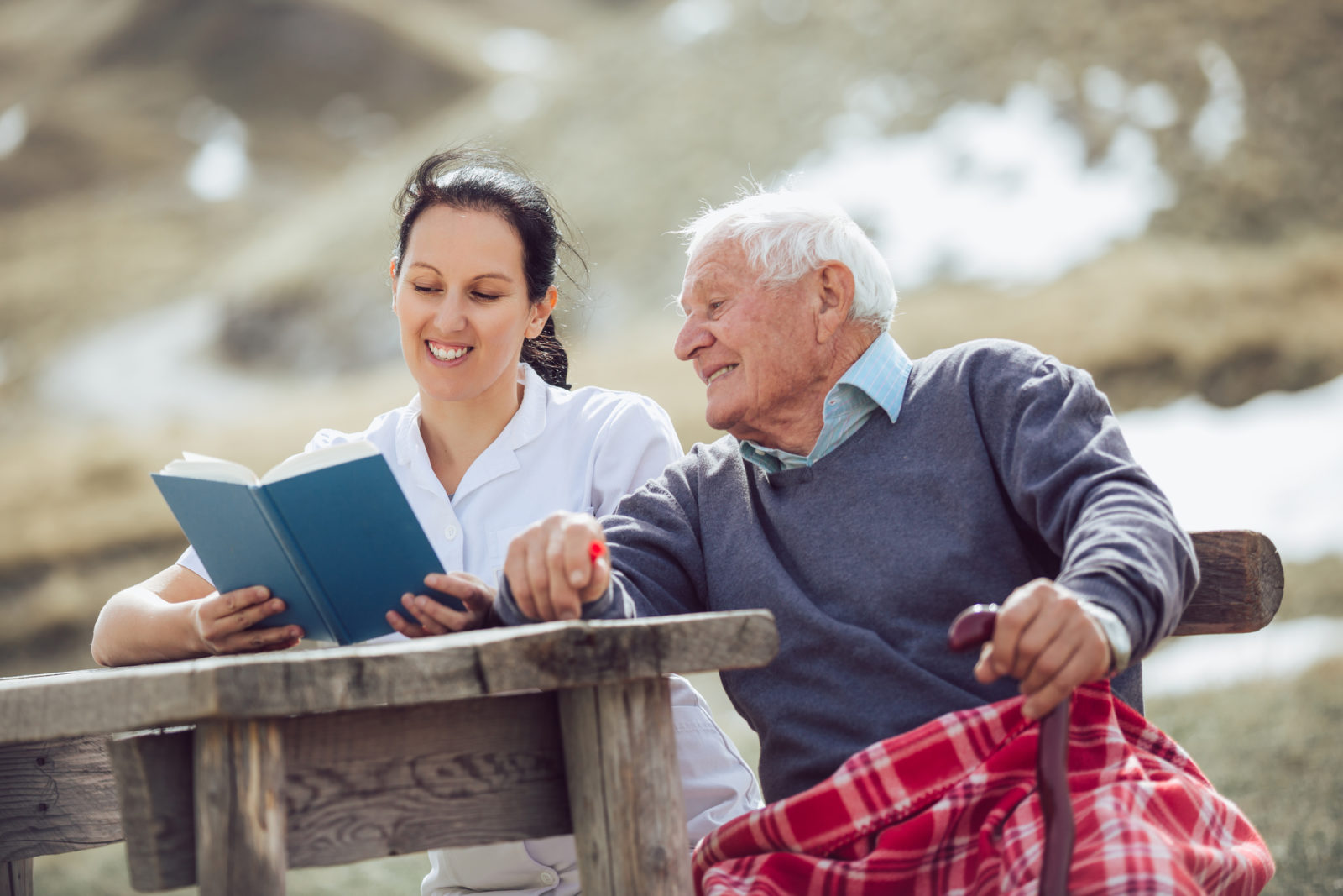 Smiling nurse reading book to gray hair senior man outdoors