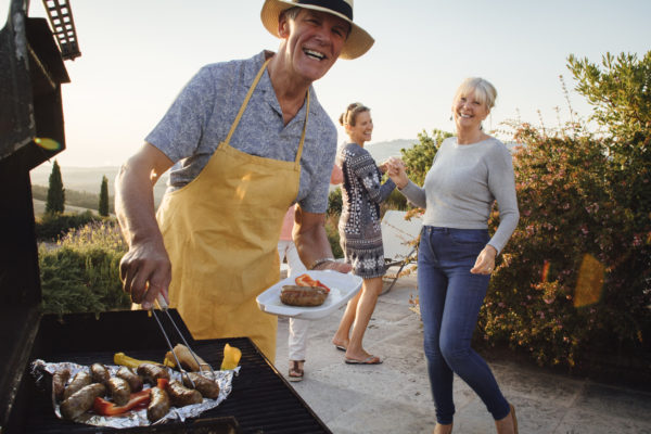 man grilling sausages outdoors, with two women in background