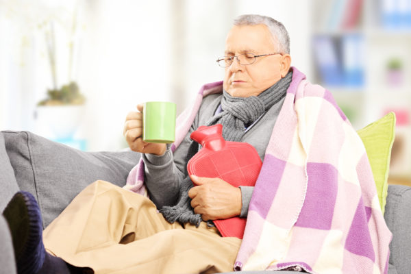 elderly man wrapped in blankets and holding a cup of hot tea and hot water bottle sitting on couch
