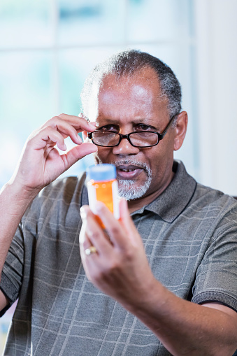 A senior African-American man in his 60s sitting at a table at home looking at the label of a bottle of prescription medicine, trying to figure out what dose to take.
