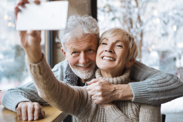 White senior couple takes a selfie inside in front of a window with a wintry outdoors