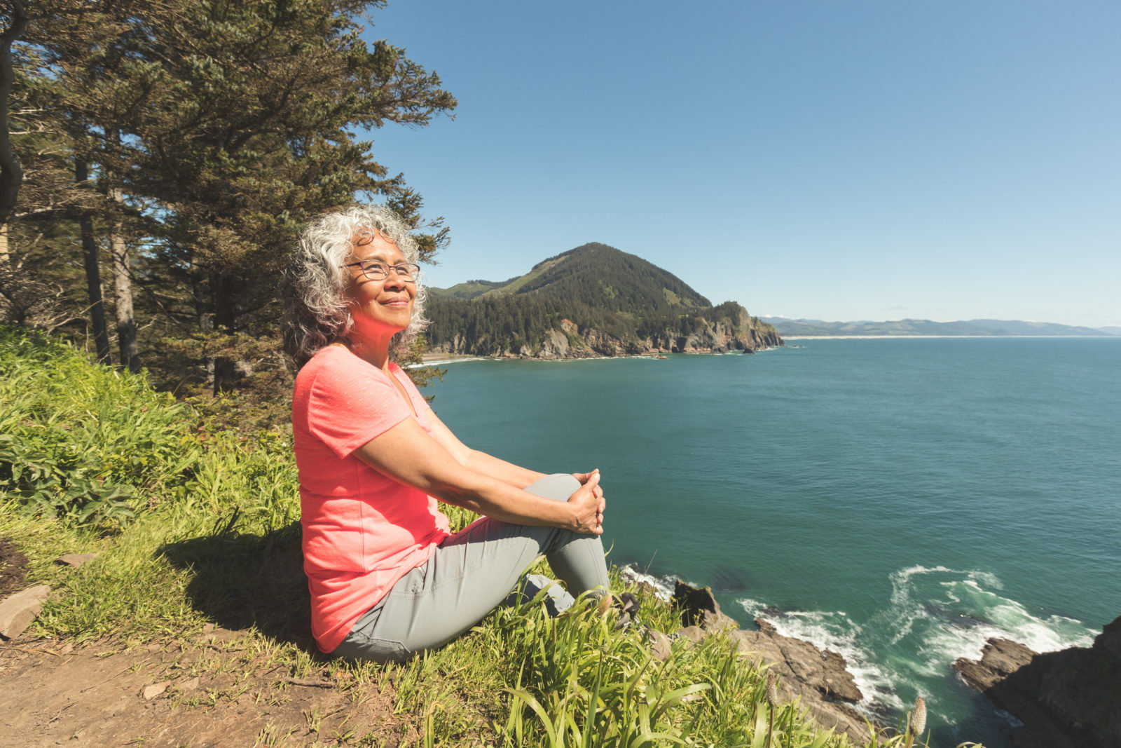 A cute senior woman relaxes on the rocks by a cliff overlooking the ocean. She has been hiking in the forest above the cliffs. A forested mountain range is in the distance and there are rocks jutting out from the ocean below.