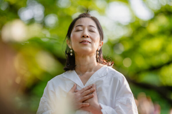 Middle aged Asian woman is closing her eyes, doing breathing exercise and meditating in nature.