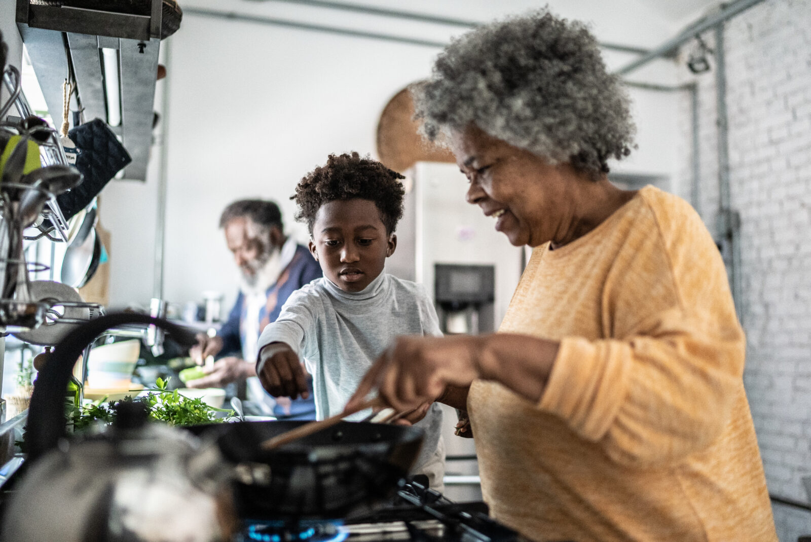 Black grandson helping his grandmother cooking at home while the grandfather looks on from the dining area.