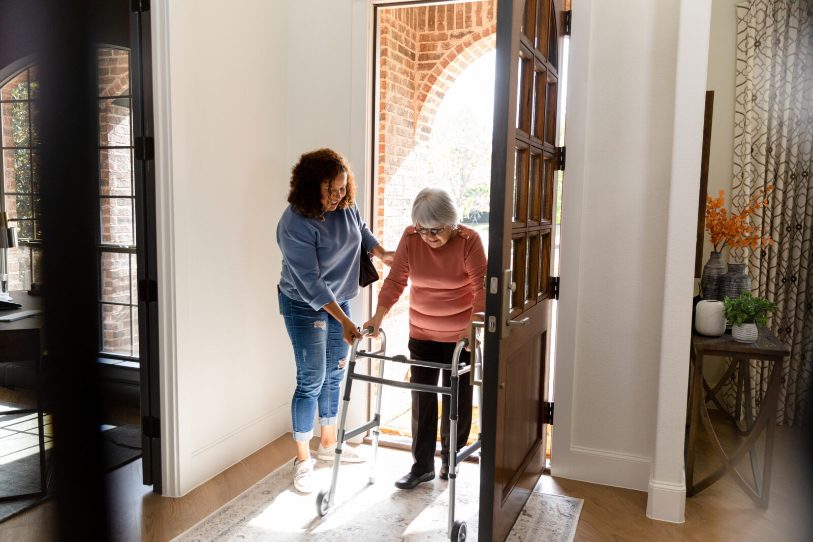 An adult woman caregiver helps a senior adult carefully enter her home.