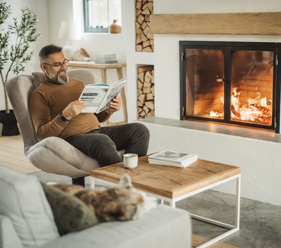 An older adult man reads the newspaper by a lit fireplace